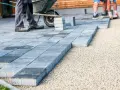Układanie płyt betonowych na podjeździe – alternatywa dla kostki. Pracownicy budowlani układają szare płyty na podsypce żwirowej.