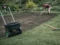 Mężczyzna zgrabia ziemię, przygotowując ją do siewu. Obok stoi wózek rozsiewający, który pomoże jak wyrównać trawnik.