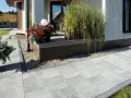 Nowy, nowoczesny chodnik przed domem z szarej kostki brukowej, ozdobiony donicami z kwiatami i trawami.