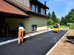 Pracownik równa łopatą świeży asfalt na podjazd, a walec go ubija. Dom w tle.