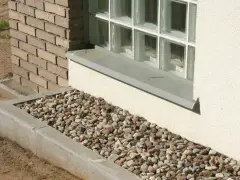 Kamienna opaska żwirowa wokół domu, otaczająca betonowy murek i okno z bloczków szklanych.