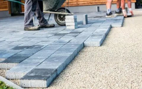 Układanie płyt betonowych na podjeździe – alternatywa dla kostki. Pracownicy budowlani układają szare płyty na podsypce żwirowej.