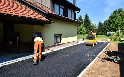 Pracownik równa łopatą świeży asfalt na podjazd, a walec go ubija. Dom w tle.