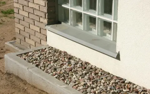 Kamienna opaska żwirowa wokół domu, otaczająca betonowy murek i okno z bloczków szklanych.