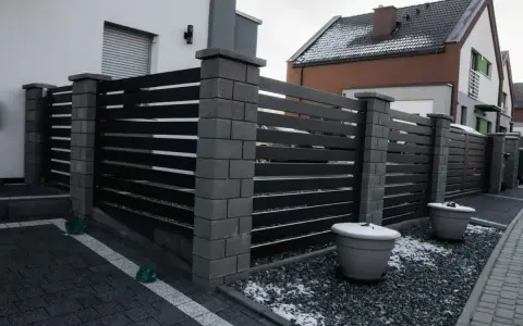 Nowoczesne ogrodzenie z betonowych słupków i ciemnych paneli. Taka wysokość ogrodzenia bez pozwolenia zapewnia prywatność.